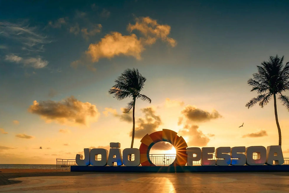 Insegna della spiaggia di JOÃO PESSOA al tramonto, con palme, nuvole dorate e oceano calmo sullo sfondo.