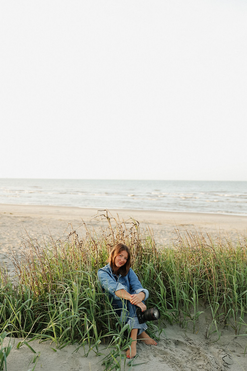 Grace sitting in beach grass with a camera, coastal dunes behind her