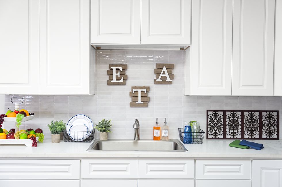 Bright white kitchen with EAT wall decor above sink, fruit tray, soap bottles, dishes, and folded towels on countertop