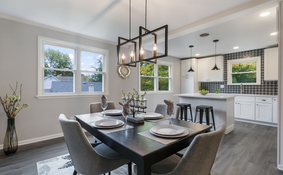 Black dining room table surrounded by light gray chairs sitting underneath a modern black chandelier with a white and black kitchen in the background