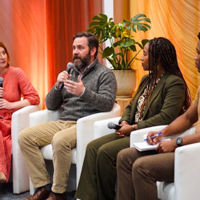 Four-panel discussion with microphones on white chairs, one man gesturing; warm orange backdrop and potted plant in a calm room.