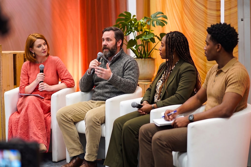 Four-panel discussion with microphones on white chairs, one man gesturing; warm orange backdrop and potted plant in a calm room.