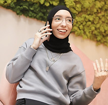 A person wearing a hijab and layered necklaces talks on the phone, with a wall of ivy visible behind them