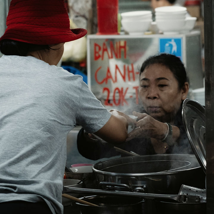 Bánh Canh, 20K — Two pairs of hands exchanging bowl of bánh canh across steaming pot at Chợ Cồn market