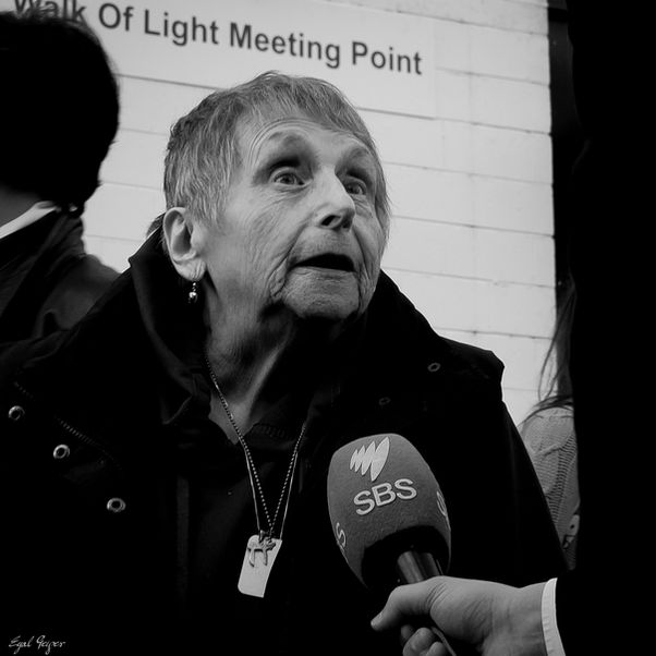 An elderly woman is interviewed by SBS News, caught mid-expression with wide eyes and open mouth, an SBS microphone held before her, a Walk of Light meeting point sign visible on the wall behind.
