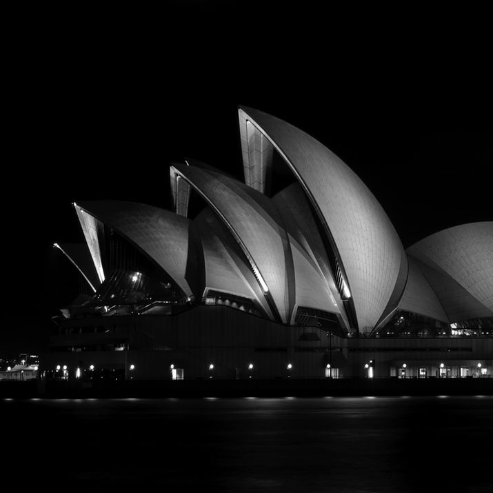 Sydney Opera House at night in black and white, the shell-shaped roof forms emerging from complete darkness against a black sky — purely sculptural, no surrounding context