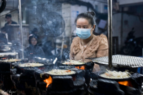 Behind the Smoke — Cook tending multiple pans over open flames through smoke at Phở Quang Gia Truyền street kitchen