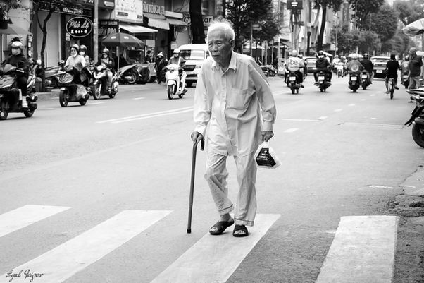 An elderly Vietnamese man in a white shirt crosses a busy Ho Chi Minh City street on a zebra crossing, walking cane in one hand and a shopping bag in the other, as streams of motorbikes flow around him.