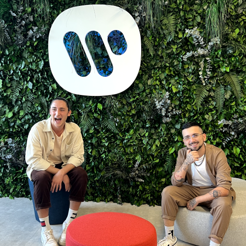 Due uomini sorridono seduti in una stanza con un muro verde dietro di loro, sul muro è il logo di Warner Music