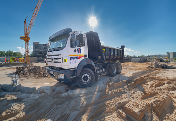 A white dump truck tilts precariously on a sandy construction site, its front wheel lodged against jagged concrete blocks. A towering crane looms in the background under a blazing midday sun, casting sharp shadows across churned earth and unfinished terrain.