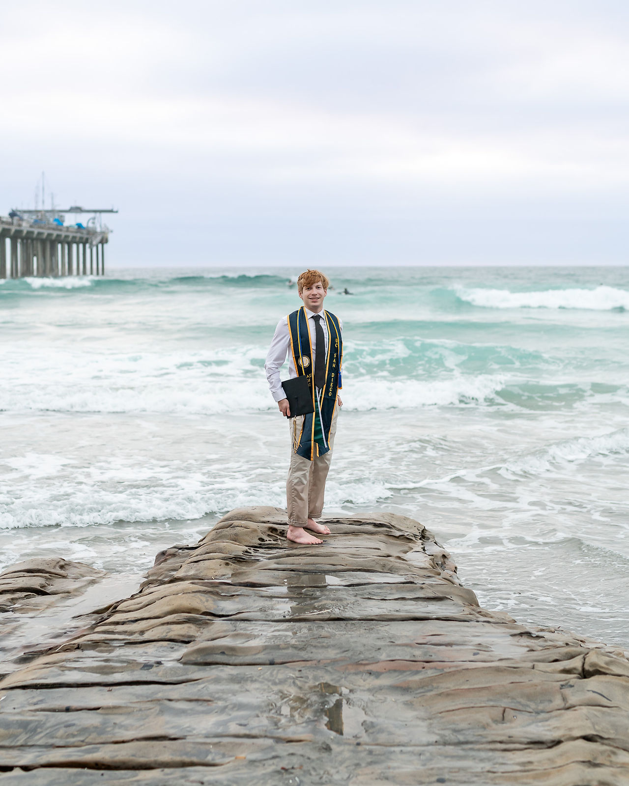 UC San Diego graduate standing barefoot under Scripps Pier in La Jolla during beach graduation photo session.