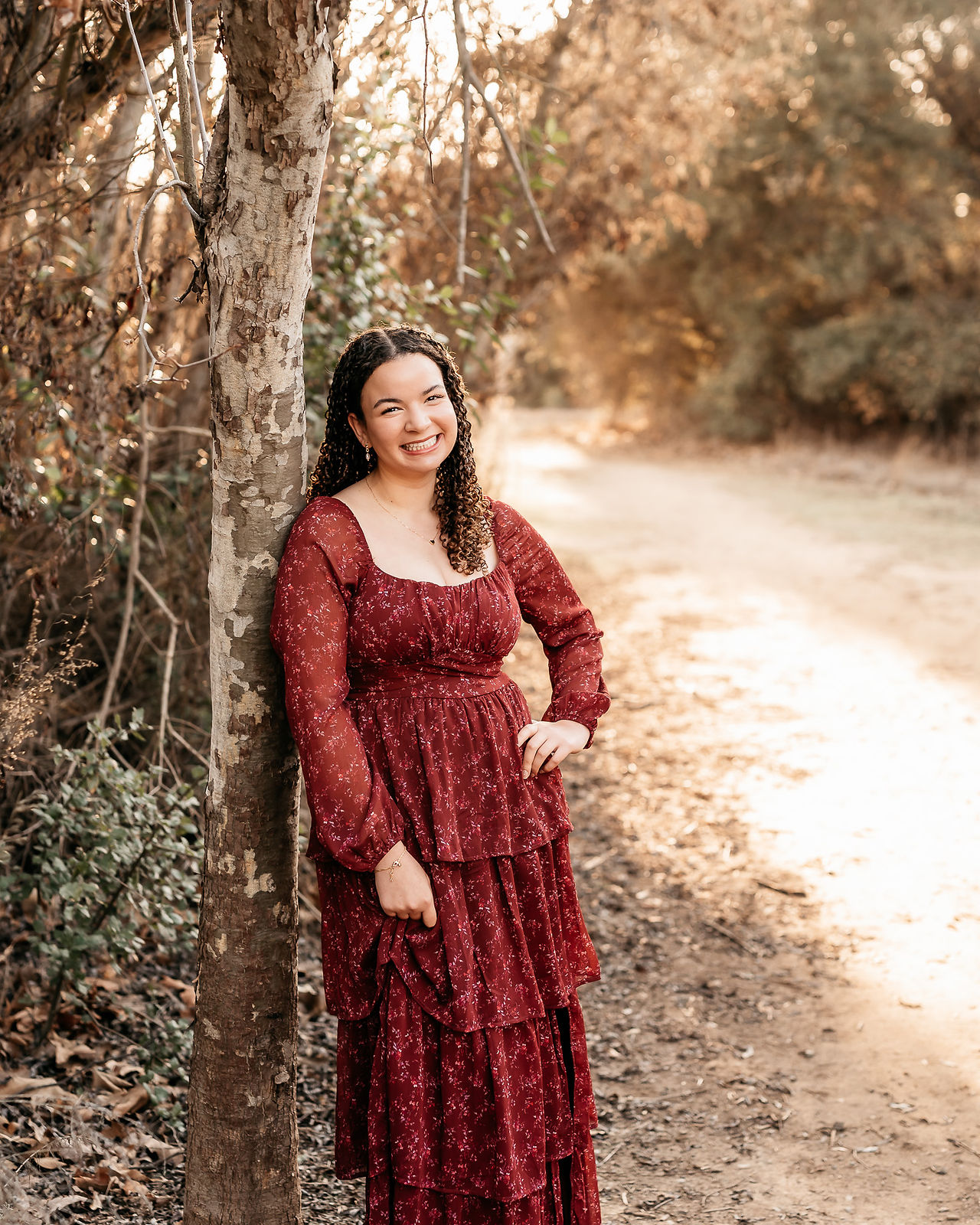 High school senior girl in a burgundy floral dress leaning against a tree on a golden San Diego trail during sunset, natural light senior portraits.