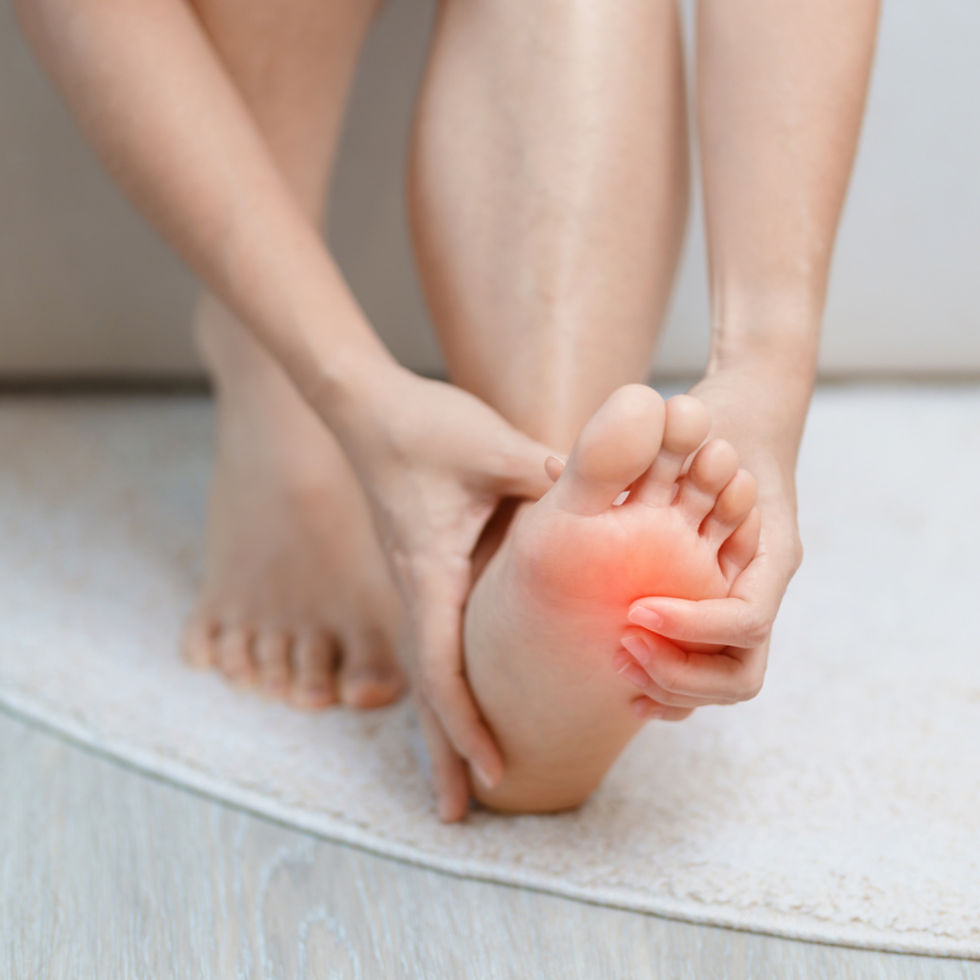 A woman sitting on a couch, reaching down and holding her foot in a slight dorsiflexion position. Red highlights the bottom of her foot, indicating pain.
