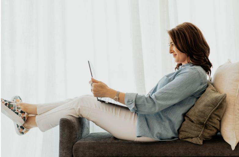 Woman in light outfit relaxing on a sofa, using a tablet. Bright room with white curtains, gray and beige cushions. Calm and focused mood.