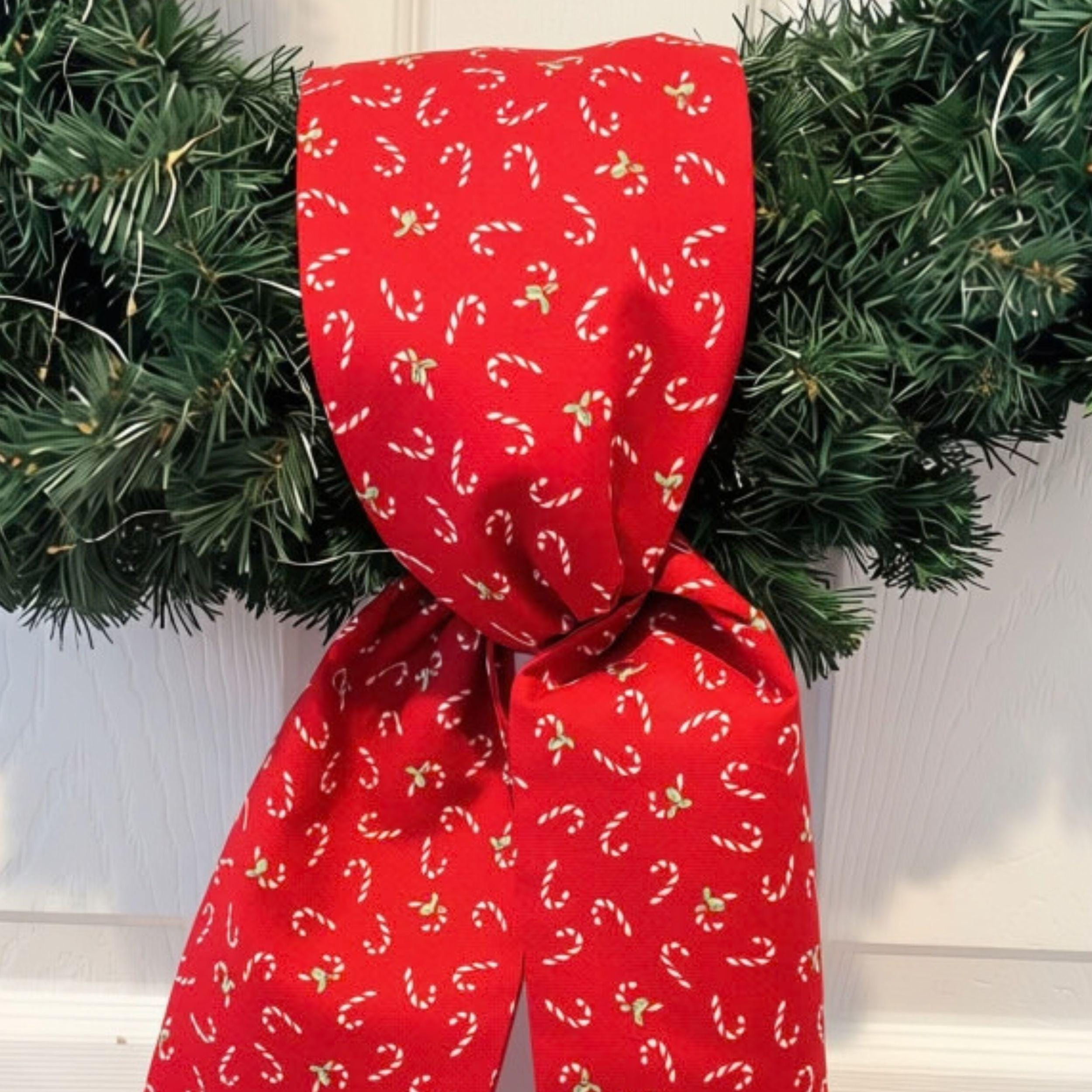 Red candy cane wreath sash displayed on evergreen wreath with warm white lights against white door