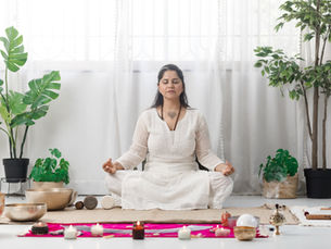 Woman in white meditating on a rug, surrounded by candles and plants. Calm, serene atmosphere with soft natural light and greenery.