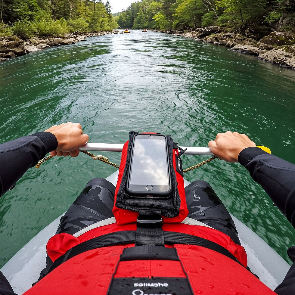 Person kayaking on a green river, holding paddles. Wearing a red life jacket with a phone in a waterproof case. Forested banks in view.