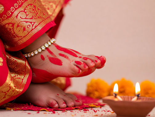 Child's feet with red patterns, anklet, in red and gold attire. Lit clay lamp and marigolds in warm setting; festive mood.
