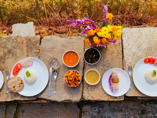Plates of rice, chapati, and vegetables on a stone slab, with vibrant flowers nearby, creating a colorful outdoor dining scene.