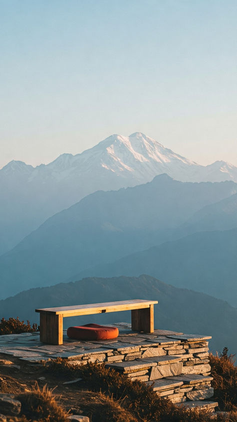 A wooden bench and red cushion on a stone platform overlook hazy mountain ranges under a clear blue sky, evoking tranquility.