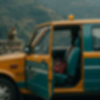 A monkey sits on a stone wall near an open teal and yellow taxi with a bag inside. Misty mountains form the background. Moody atmosphere.