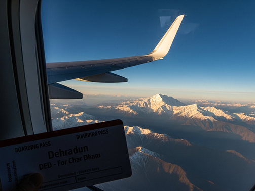 Plane window view of snow-capped mountains at sunset. A boarding pass for Dehradun-Char Dham is visible, creating a serene travel mood.