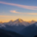 Snow-capped mountains under a vibrant sky with swirling clouds at sunrise or sunset, casting a warm glow. Peaceful and majestic scene.