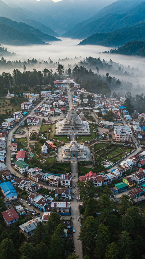Aerial view of a temple complex surrounded by houses in a misty valley, framed by dense green forest and distant mountains. Peaceful mood.