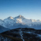 Snow-capped mountain peaks under a clear blue sky, with a winding path on a dark hill in the foreground. Peaceful and majestic scene.