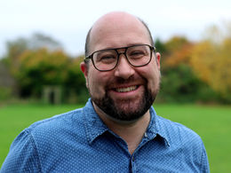 Alex, a white bearded man with a bald head, glasses and wearing a blue shirt, is outdoors and smiling at the camera