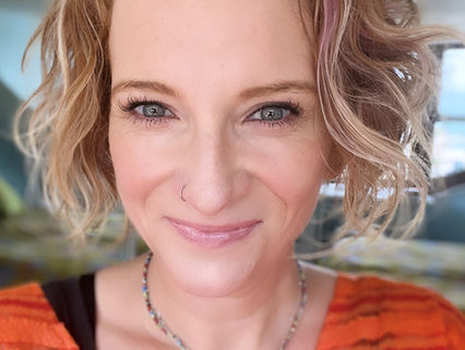 Abi, a white woman with short, blonde, wavy hair and wearing an orange top is smiling at the camera