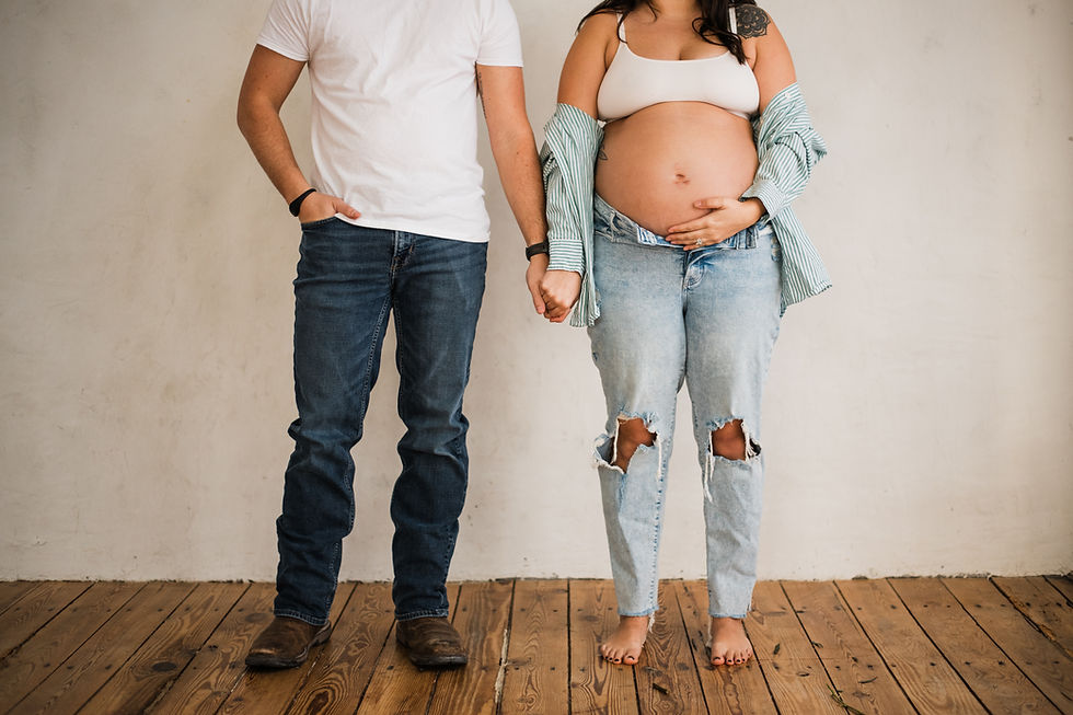 Pregnant woman in ripped jeans and striped shirt holds belly, stands barefoot with a man in a white shirt and jeans, holding hands on wood floor.