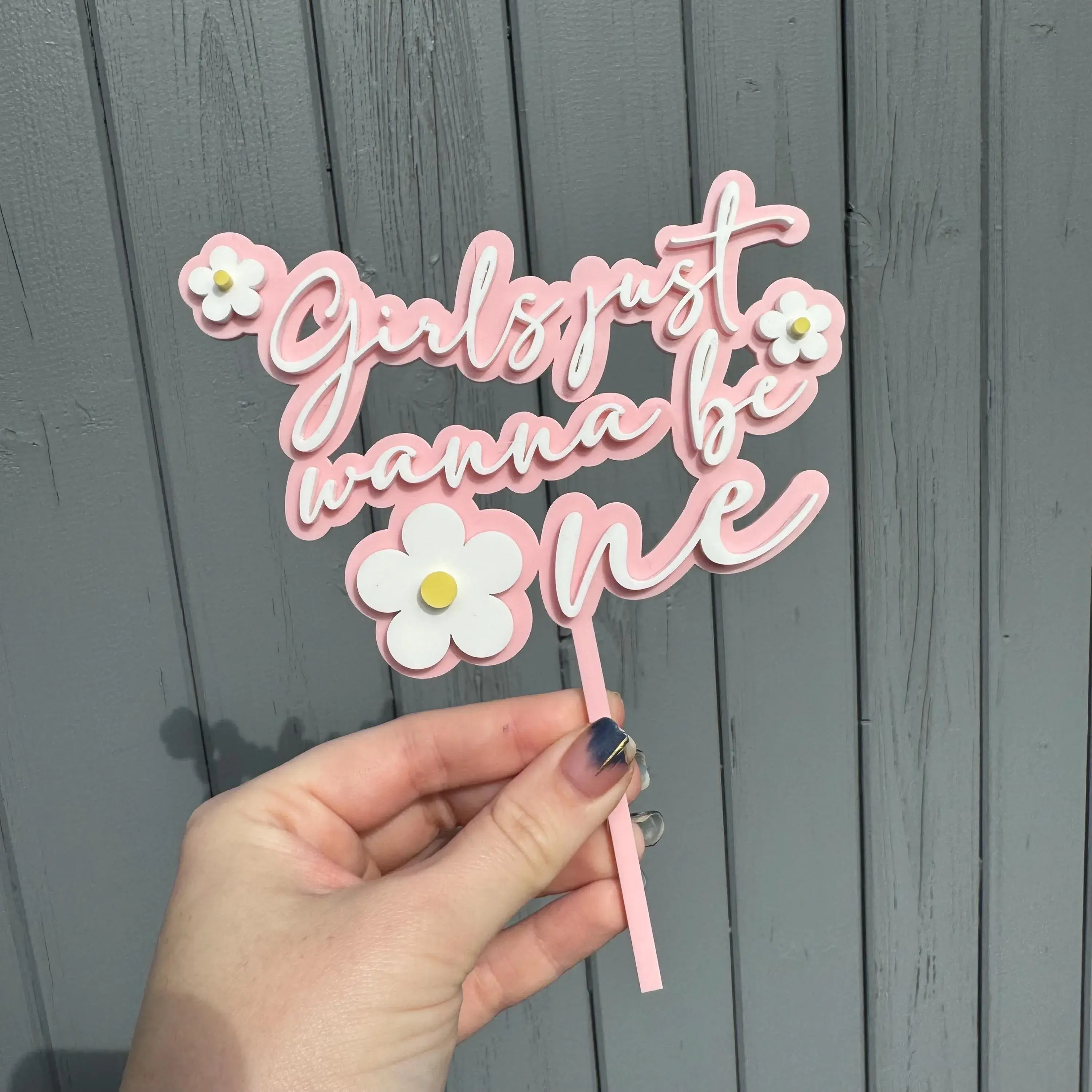 Hand holding pink and white cake topper with text Girls just wanna be and daisies
