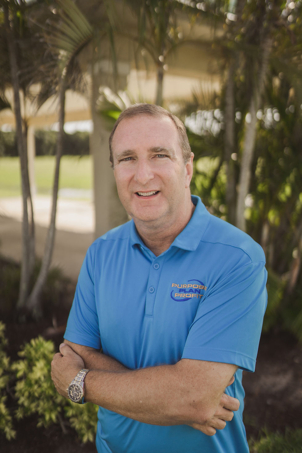man with grey hair and blue polo shirt with arms cross on wooded outdoors background