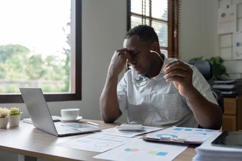 Man sitting at a desk, holding glasses, looking stressed. Papers and a laptop are in front of him, with a window view of greenery. He's overwhelmed by amount of time it takes updating QuickBooks Online cost and prices.