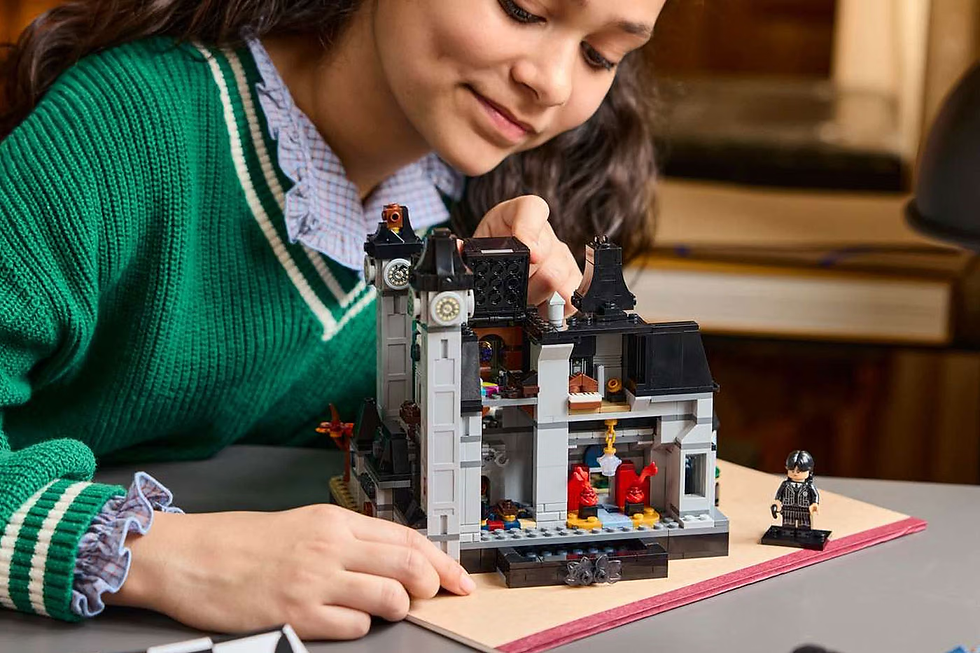 Yeşil hırkalı bir kız, masada LEGO ev modeli kuruyor; yanında küçük figür var, iç mekânda odaklı ve mutlu.
