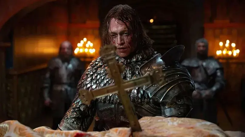 Zırhlı bir şövalye, mumlarla aydınlatılmış karanlık bir odada haça bakarken duruyor. Arka planda başka zırhlı askerler var. Dram dolu atmosfer.