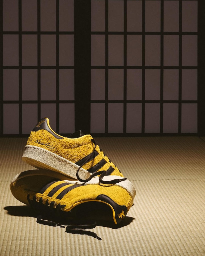 Sarı renkli spor ayakkabılar, tatami zemin üzerinde üst üste duruyor. Arkadaki siyah-beyaz panel, minimalist bir ortam sunuyor.