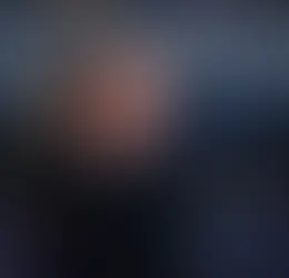 Sahnedeki kişi, siyah ceketli, kollarını açmış ve mikrofona doğru bakıyor. Arkada bulanık bir kalabalık var, sahne ışıkları parlıyor.