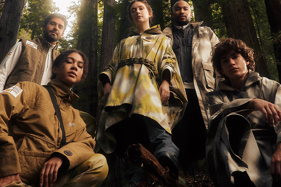 Beş kişi ormanda poz veriyor, koyu tonlarda ve desenli kıyafetler giymişler. Arka planda dev ağaçlar var. Ciddi yüz ifadeleri.