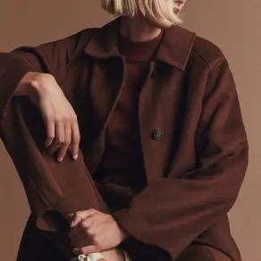 Blond bir kişi, kahverengi bir kaban ve pantolon giyiyor, beyaz ve kahverengi spor ayakkabılı. Bej arka plan, rahat bir pozda oturuyor.