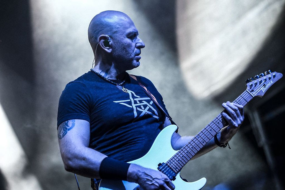 Kel bir adam gitar çalıyor, siyah bir tişört giyiyor, üzerinde beyaz bir sembol var. Sahne ışıkları altında odaklanmış görünüyor.