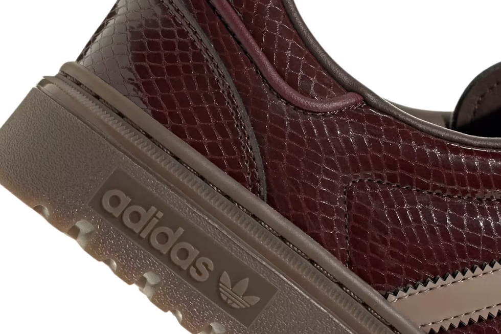 Kahverengi yılan derisi desenli ayakkabı detayı. Üstte "adidas" metni ve markanın logosu var. Arka plan beyaz.