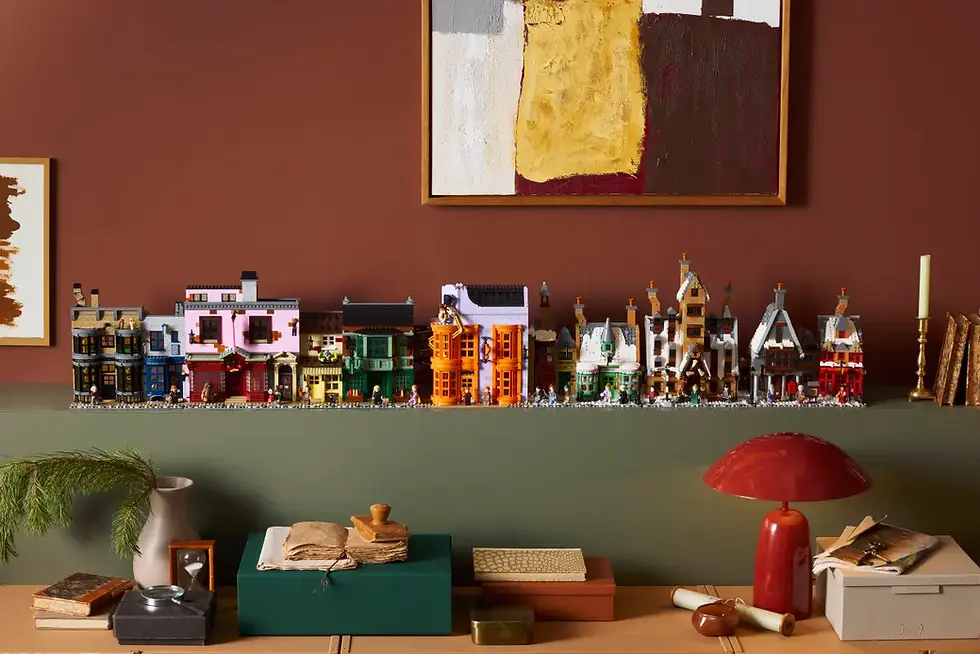 Mini Lego binaları, yeşil ve kahverengi duvar önünde, masa üstünde sergileniyor. Masada kitaplar ve kırmızı lamba var. Sanat eseri duvarda.