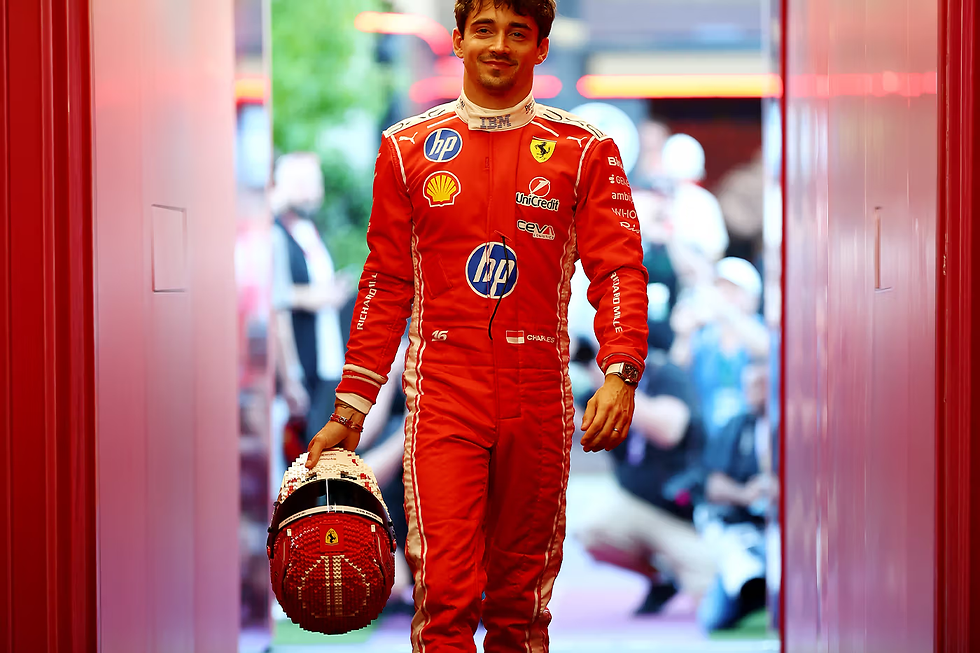 Kırmızı yarış tulumlu bir adam, kaskını tutarak gülümsüyor. Arka planda bulanık izleyiciler ve ışıklar var. Yarış pisti ortamı.