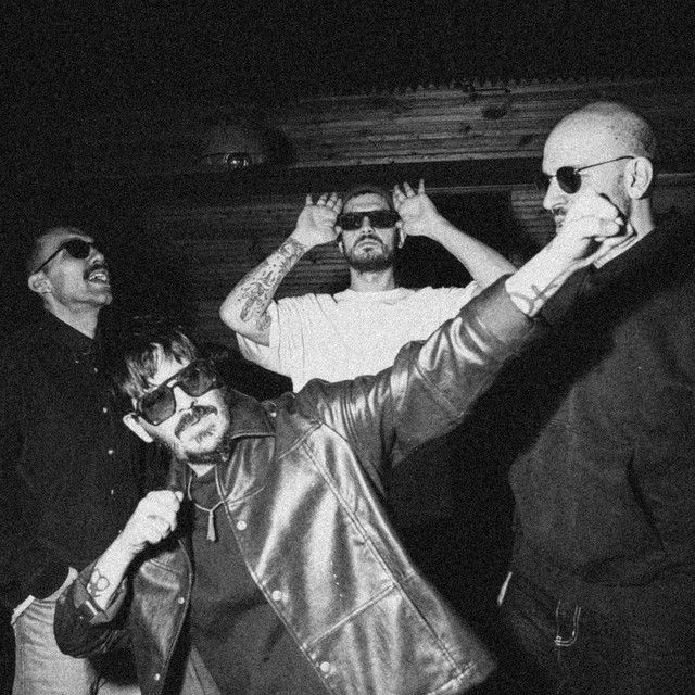 Siyah beyaz fotoğrafta, güneş gözlüklü dört adam, eğlenceli pozlar veriyor. Ahşap arka plan, rahat bir atmosfer yaratıyor.
