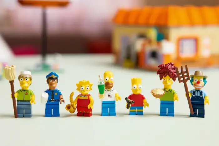 Yedi LEGO figürü yan yana dizilmiş. Arka planda bulanık bir yapı var. Figürler canlı renklere sahip, bazıları aksesuar tutuyor.