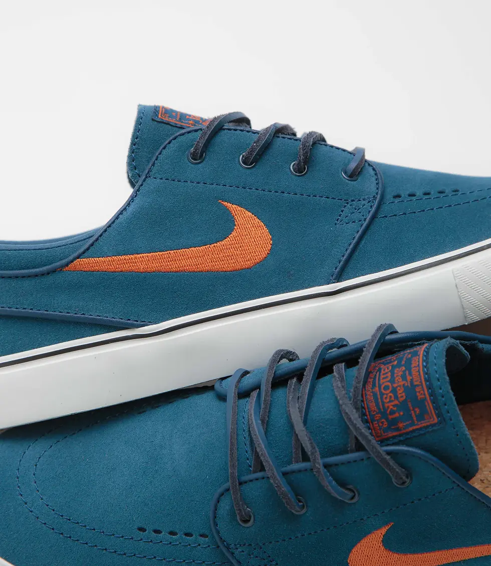 Mavi sneaker ayakkabılar beyaz tabanlı, turuncu logo ve bağcıklarla. Üzerinde turuncu etikette yazı görülüyor. Arka plan beyaz.