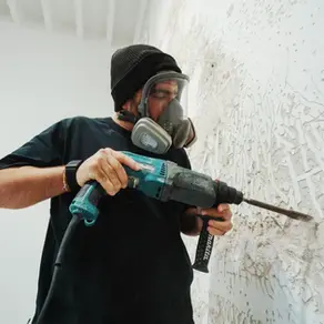 Bir kişi, siyah şapka ve gaz maskesi takarak beyaz duvarda elektrikli matkapla çalışıyor. Arka plan beyaz, duvar dokulu.