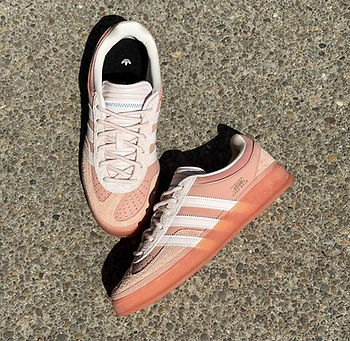 Pembe-beyaz spor ayakkabılar, taşlı zeminde sergilendi. Ayakkabı üzerinde beyaz şeritler ve Adidas logosu dikkat çekiyor.